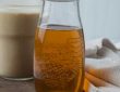 A clear glass bottle of homemade sugar-free vanilla syrup with allulose, sitting next to a steaming latte