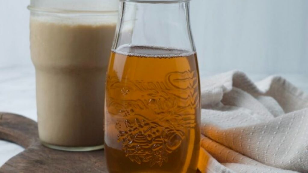 A clear glass bottle of homemade sugar-free vanilla syrup with allulose, sitting next to a steaming latte
