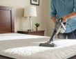 A person using a handheld vacuum cleaner with an upholstery attachment to deep clean a white mattress in a bright bedroom