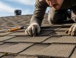 A roofing contractor performing a close-up inspection of asphalt shingles on a residential roof, checking for cracks and missing sections during a routine maintenance visit.