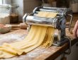 Stainless steel manual pasta machine with hand crank on a wooden kitchen countertop