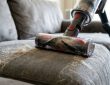 A person using a handheld vacuum with an upholstery attachment to remove pet hair from a gray fabric couch
