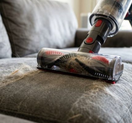 A person using a handheld vacuum with an upholstery attachment to remove pet hair from a gray fabric couch